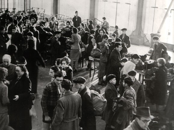 Black and white photograph of a large hall crowded with refugees, many of them young people and children, standing and talking.