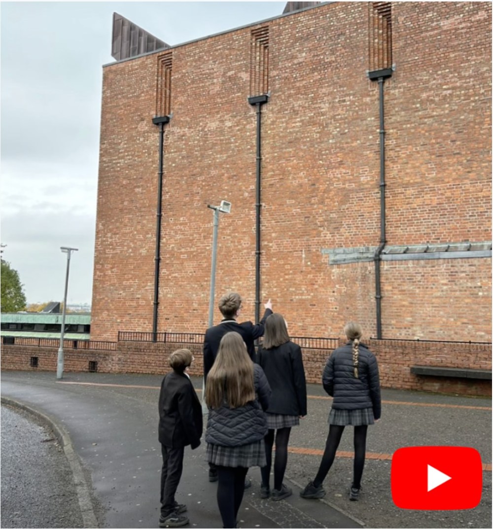 Four pupils standing on a pavement looking up at the tall red brick wall of a church, one pointing upwards. A YouTube play button is in the corner.