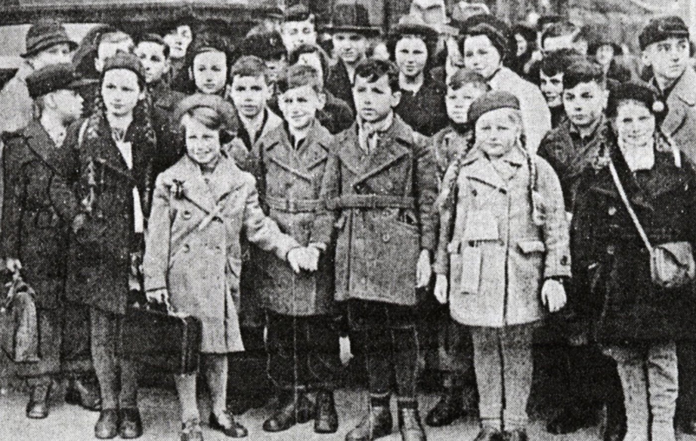 Black and white photograph of a large group of children in coats and hats, several holding suitcases, standing close together outdoors.