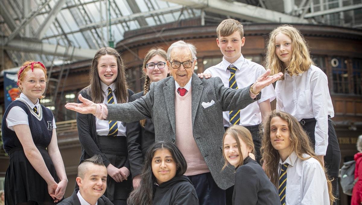 An elderly man with arms outstretched, surrounded by smiling school pupils in a railway station.
