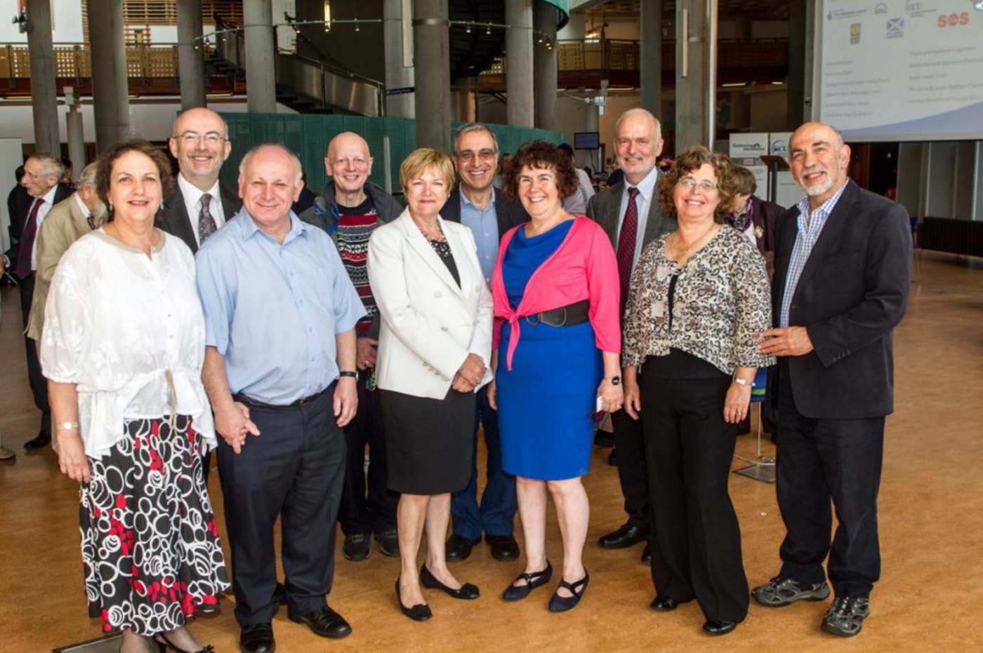Nine adults standing together in a large hall, smiling at the camera: the volunteers, a university principal and guests at a Gathering the Voices event in 2013.