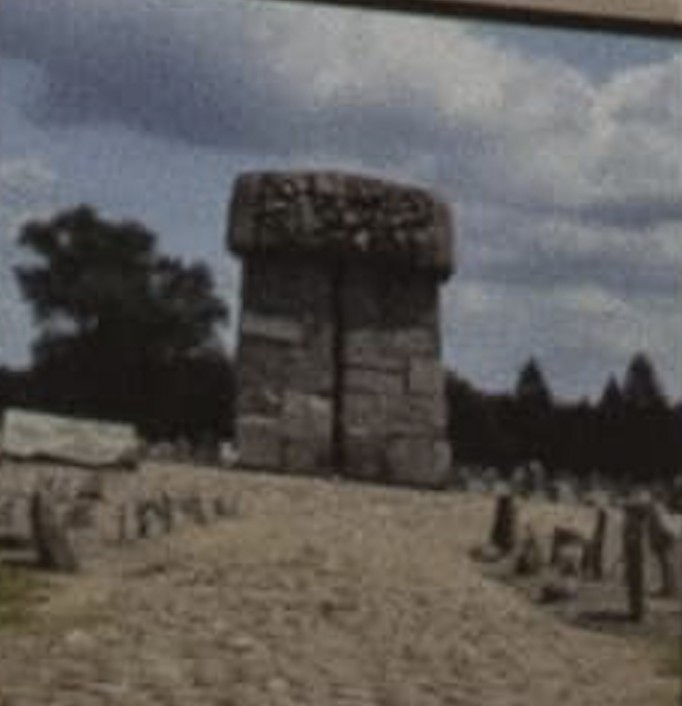 The memorial monument at Treblinka, a tall stone block above a field of upright stones, under a cloudy sky.