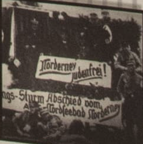 Black and white photograph of a parade float draped with a banner reading Norderney judenfrei, with uniformed men standing on it.