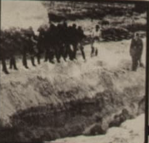 Black and white photograph of a line of people standing beside a large pit in the snow.