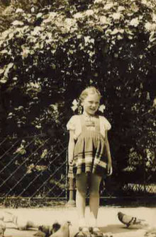 Sepia photograph of a smiling young girl in a checked dress standing in front of a hedge, with pigeons at her feet.
